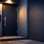 Modern front door entrance at dusk, sleek smart doorbell camera mounted beside a dark charcoal door, soft porch lighting casting warm glow against cool evening tones, shallow depth of field, clean architectural lines, slate blue and deep charcoal color palette with soft white accents