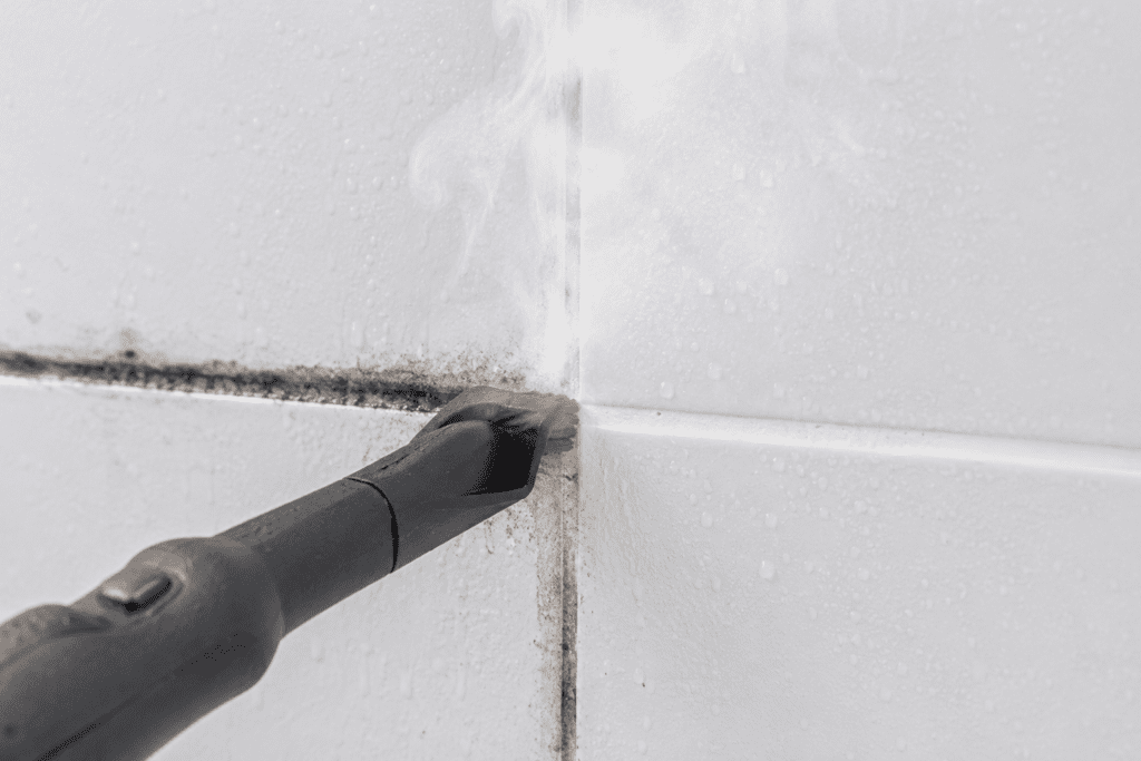 Close-up of a steam cleaner nozzle pushing steam into white tile grout lines in a clean modern bathroom. Water vapor visible, bright lighting, satisfying before/after visual contrast between a clean and grimy grout line. Photorealistic macro-style shot.
