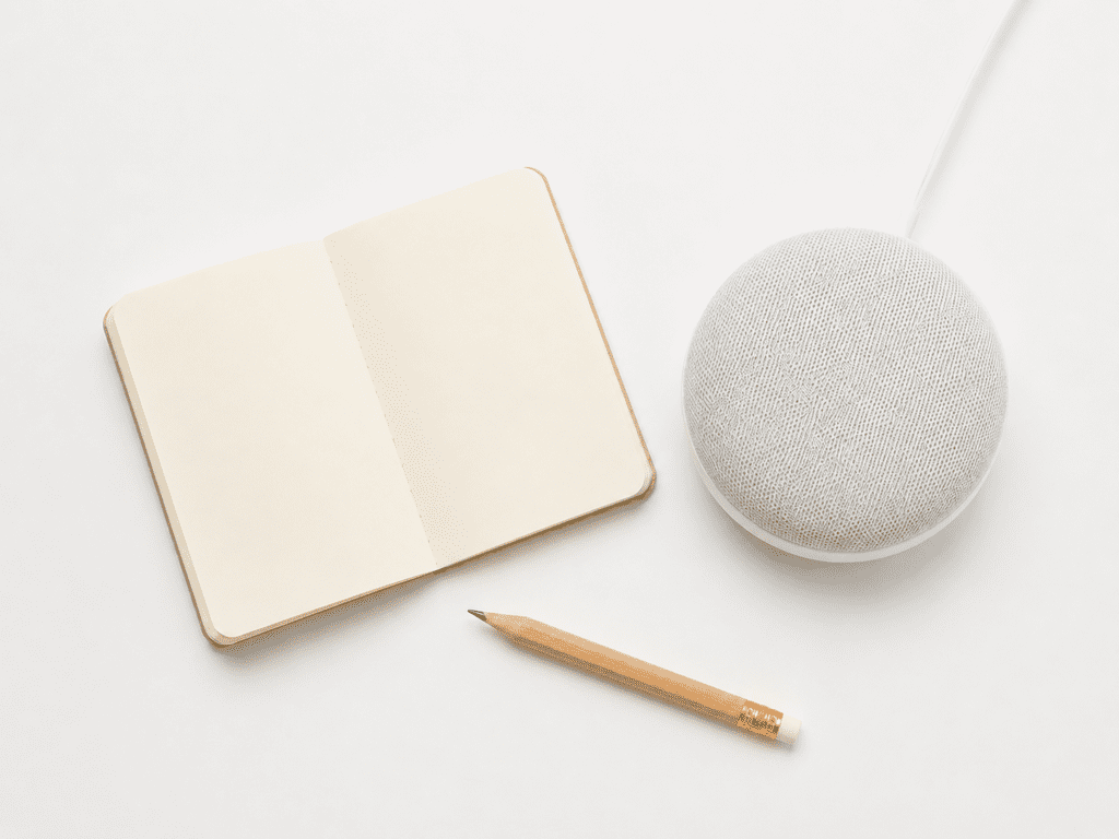 Flat lay of a small open notebook, a short pencil, and a single modern smart home device a compact white smart speaker arranged simply on a clean white surface with soft natural light. Minimal styling, generous negative space, no hands, no people, no text. Bright white background, slightly warm tone. Shot from directly above. Product photography aesthetic, crisp and clean.