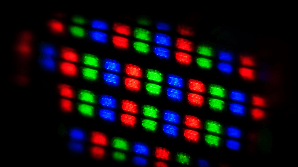 Close-up macro shot of a single illuminated screen pixel cluster glowing in red, green, and blue against a deep black background. Light diffuses softly outward from each colour point. Dark, dramatic, cinematic.