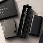 Neatly arranged Father's Day gift flatlay on a light grey linen surface, featuring a slim metal wallet, a multitool pen in a gift box, a compact power bank, and small cologne sample vials, warm natural window light, styled like a lifestyle magazine, top-down angle.