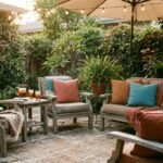 A cozy backyard patio at golden hour, warm string lights overhead, comfortable outdoor seating with cushions in terracotta and warm teal tones, a small side table with drinks, a shade umbrella, lush greenery in the background, inviting and lived-in atmosphere, photorealistic lifestyle photography, soft warm evening light