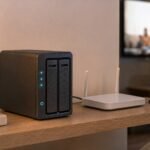 A compact two-bay home NAS with glowing status lights on a living room shelf, wired to a white router, with a smartphone beside it and a TV and laptop in the background.